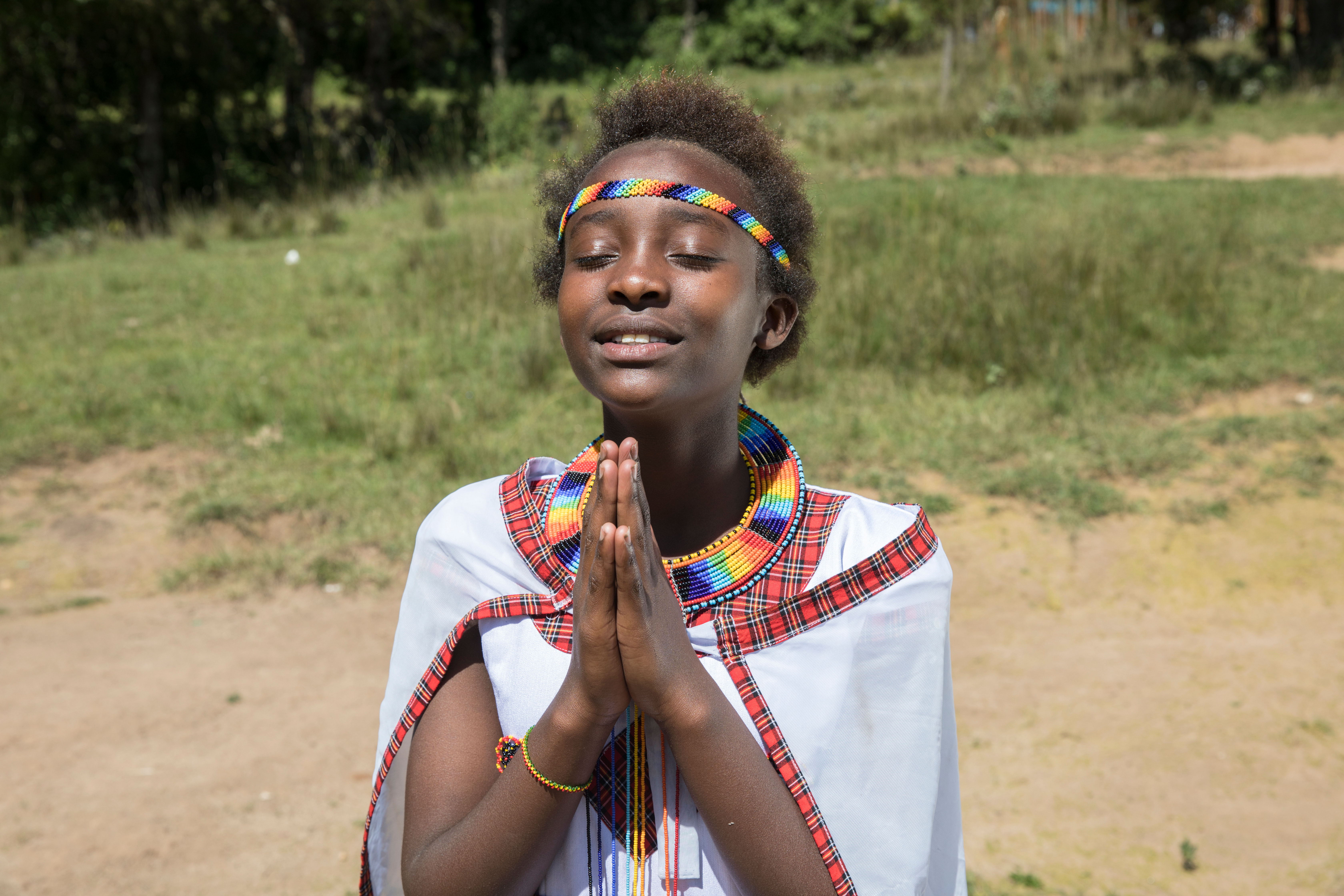 Young girl wearing a bright beaded headband and white dress clasps her hands together with her eyes closed asa she prays.