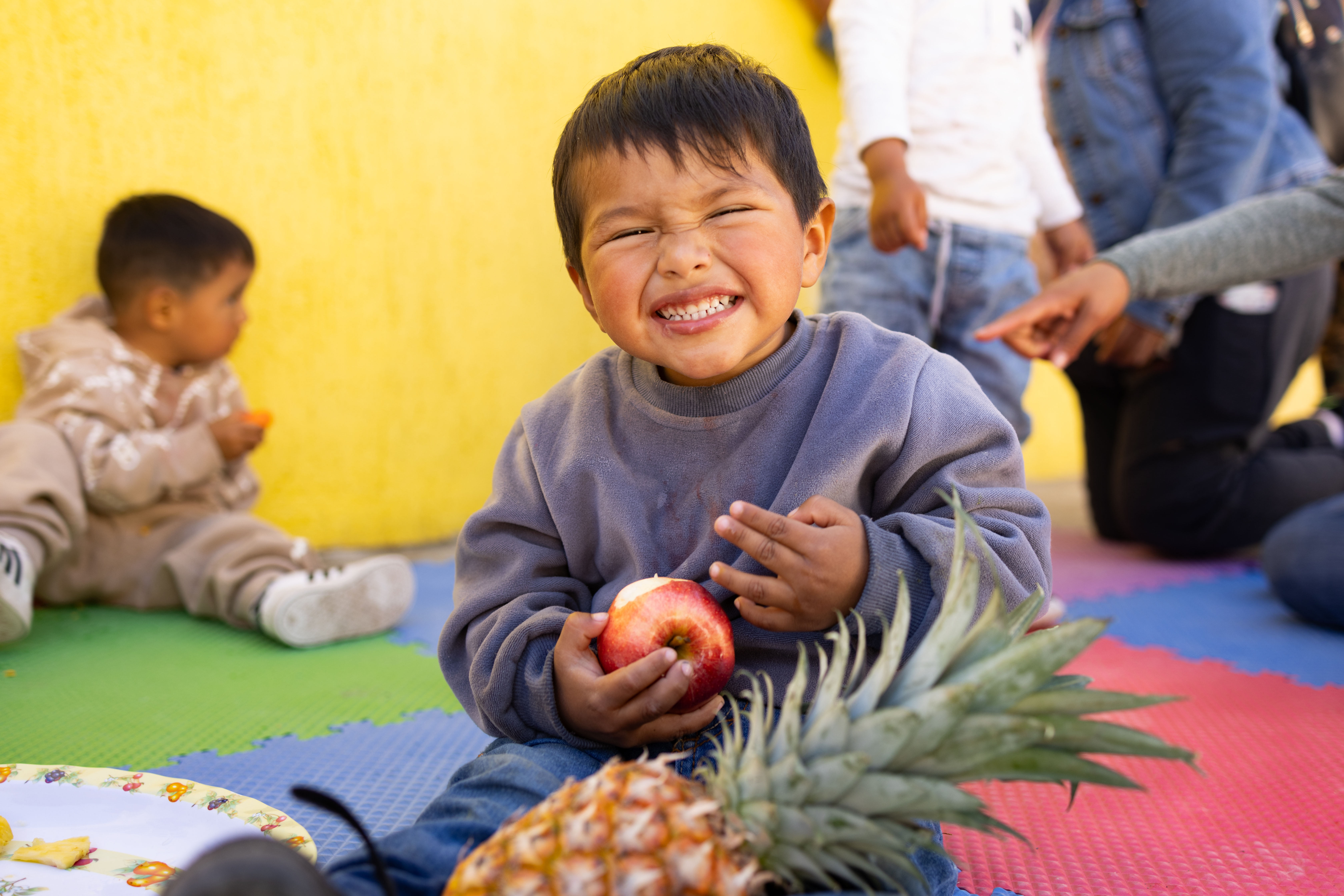 A toddler smiles for the camera while holding an apple and a pineapple.