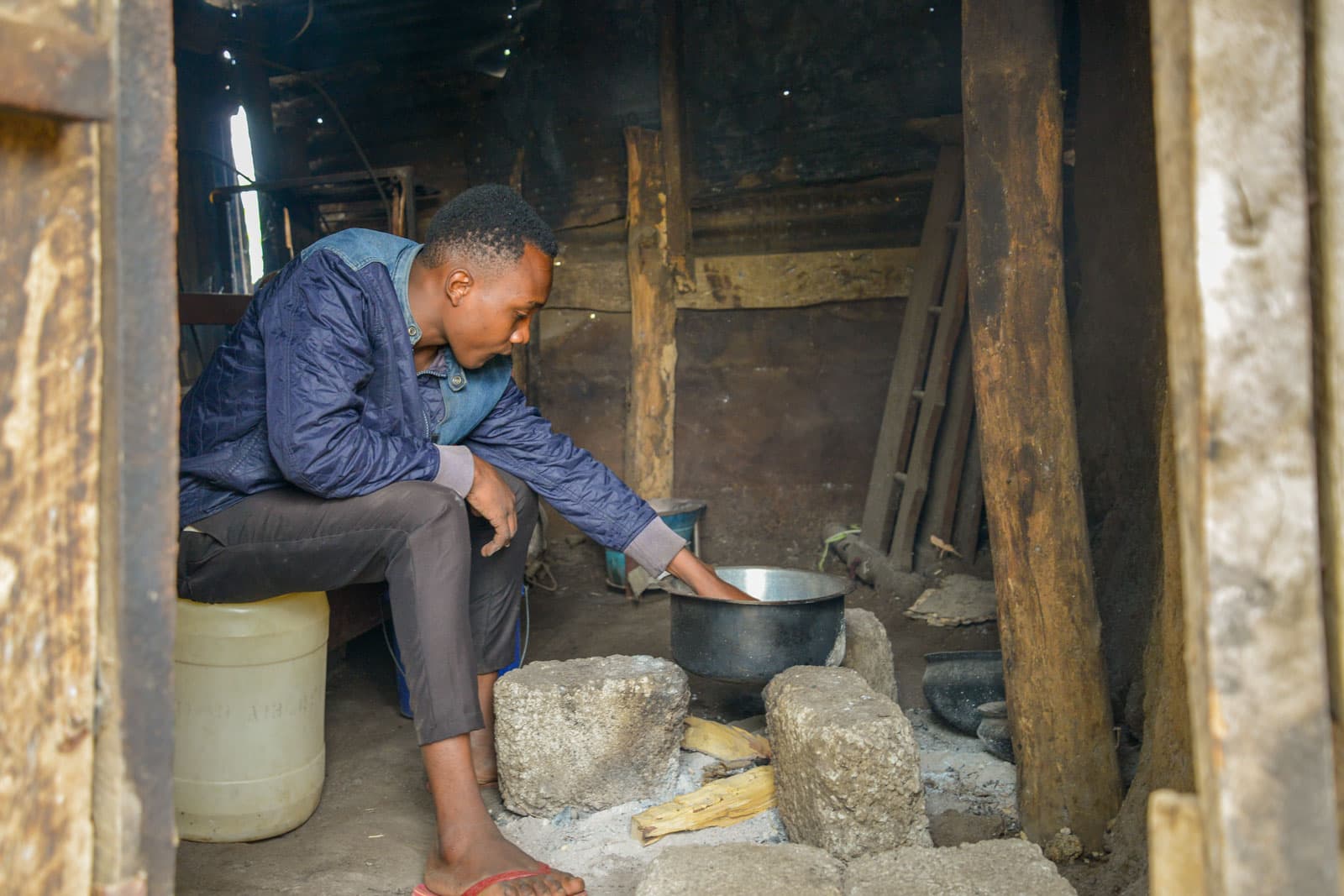 Moses is wearing a blue jacket and gray pants. He is roasting peanuts in a pot inside his kitchen.