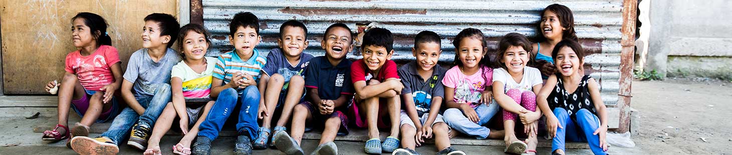 Many children sitting on the ground together with their backs leaning against a wall