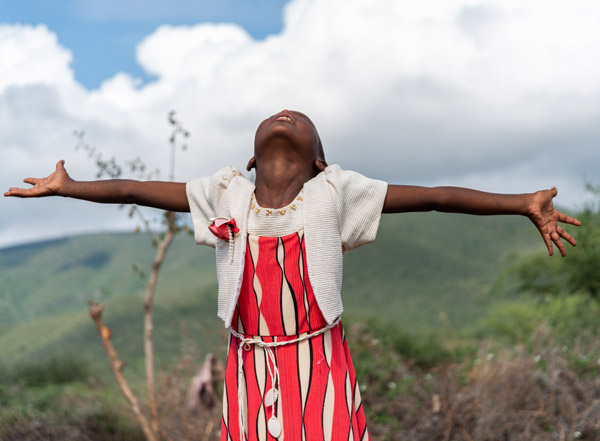 Young girl wearing a pattered red and cream dress lifts her head and raises her arms to the sky.