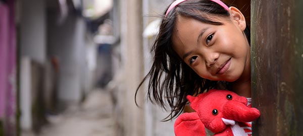 A girl holding a red stuffed elephant peaks her head out from behind a pole