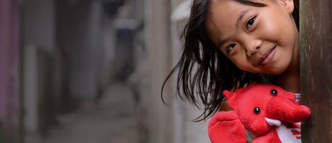 A girl holding a red stuffed elephant peaks her head out from behind a pole