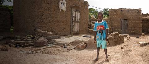 A barefoot boy in a blue t-shirt and blue shorts stands in front of a mud house