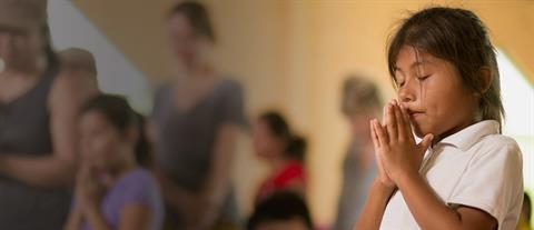 A young girl in a white shirt with her eyes closed and her hands together in prayer