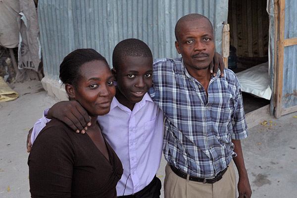 A boy stands outside his home with arms around his mother and father