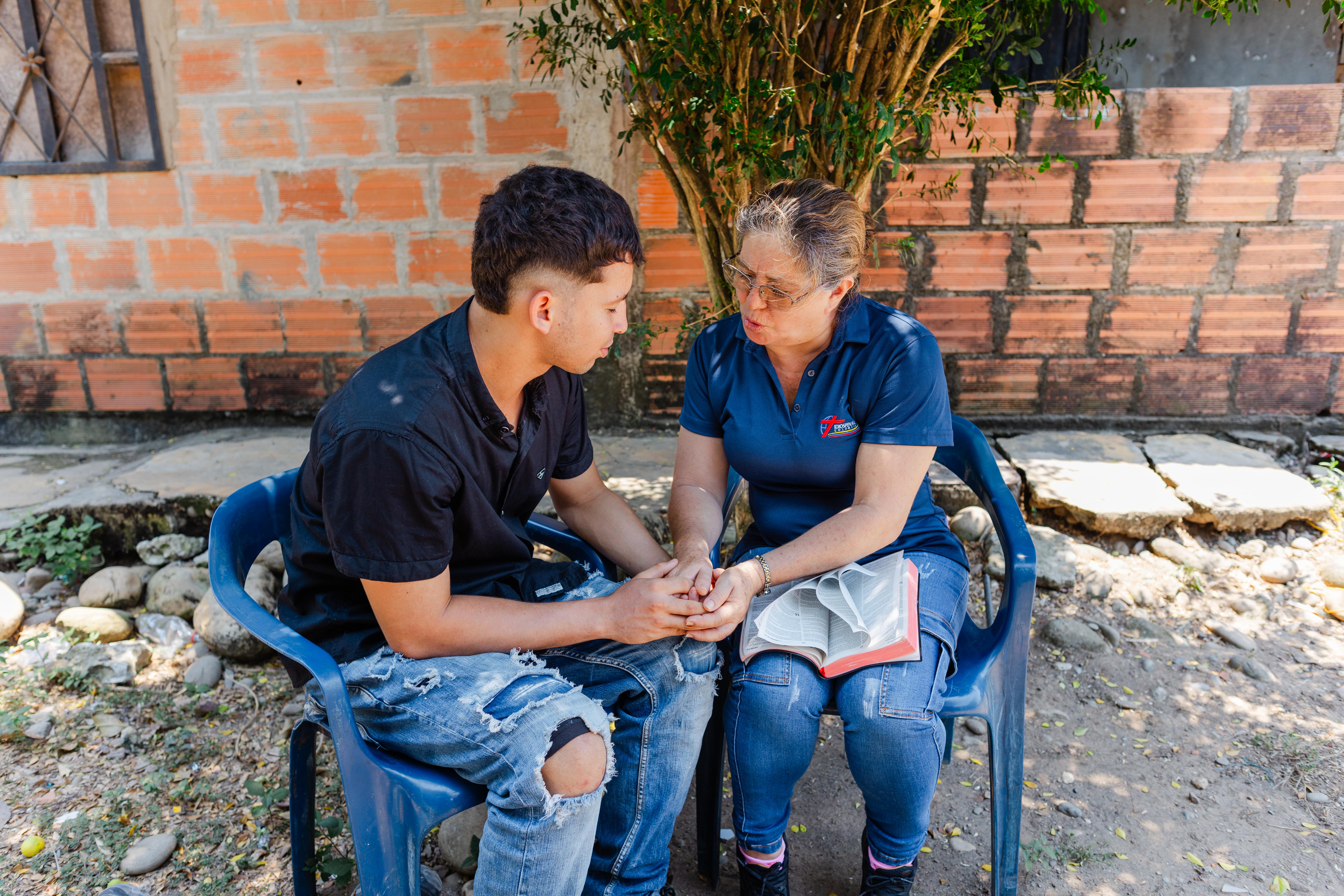 A woman with a Bible in her lap holds the hands of a teenage boy as they both pray.