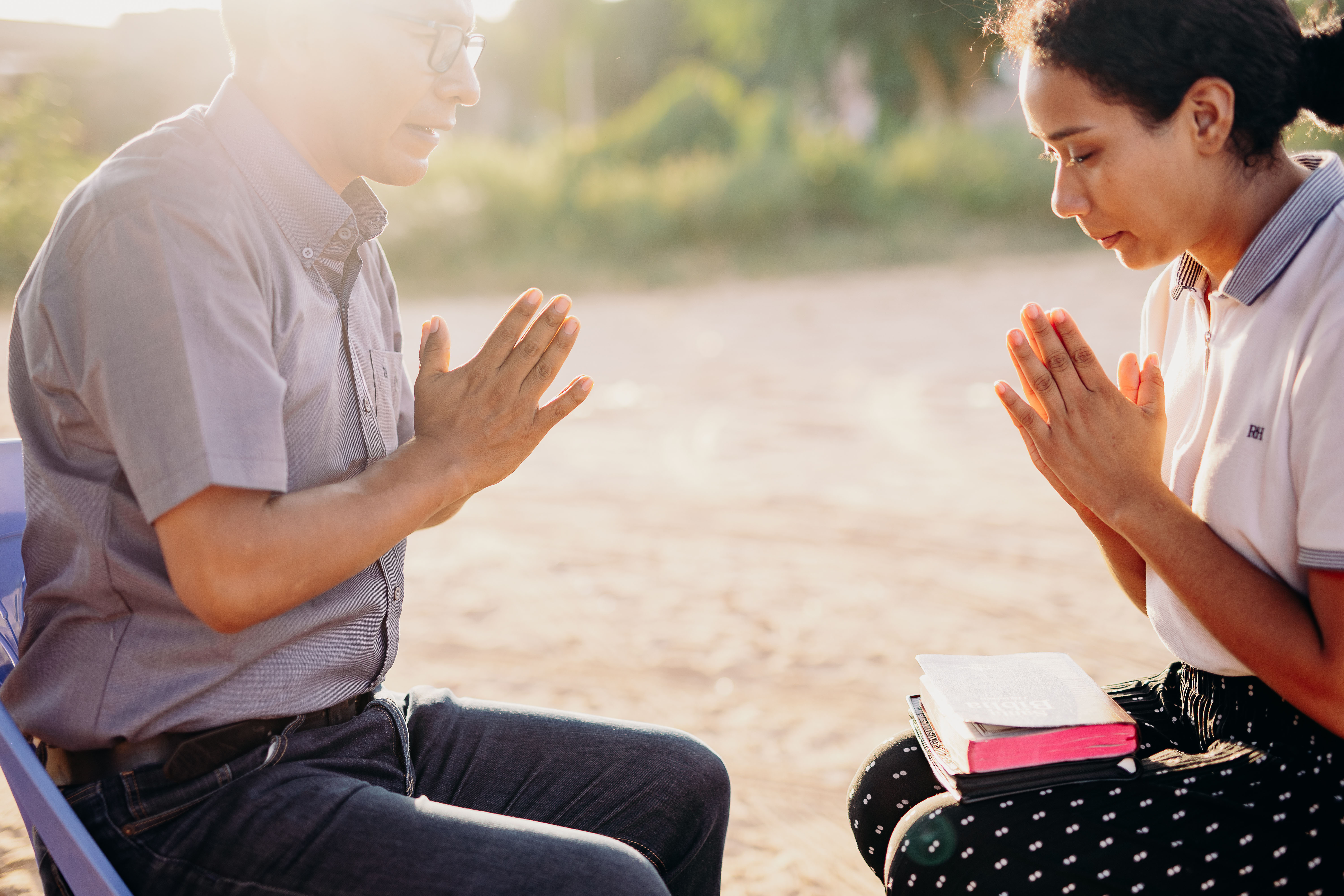 Young woman learns how to pray by sitting across from her pastor as bright light shines in the top left corner. Both the woman and her pastor have their hands placed together in front of their chests and their eyes closed.