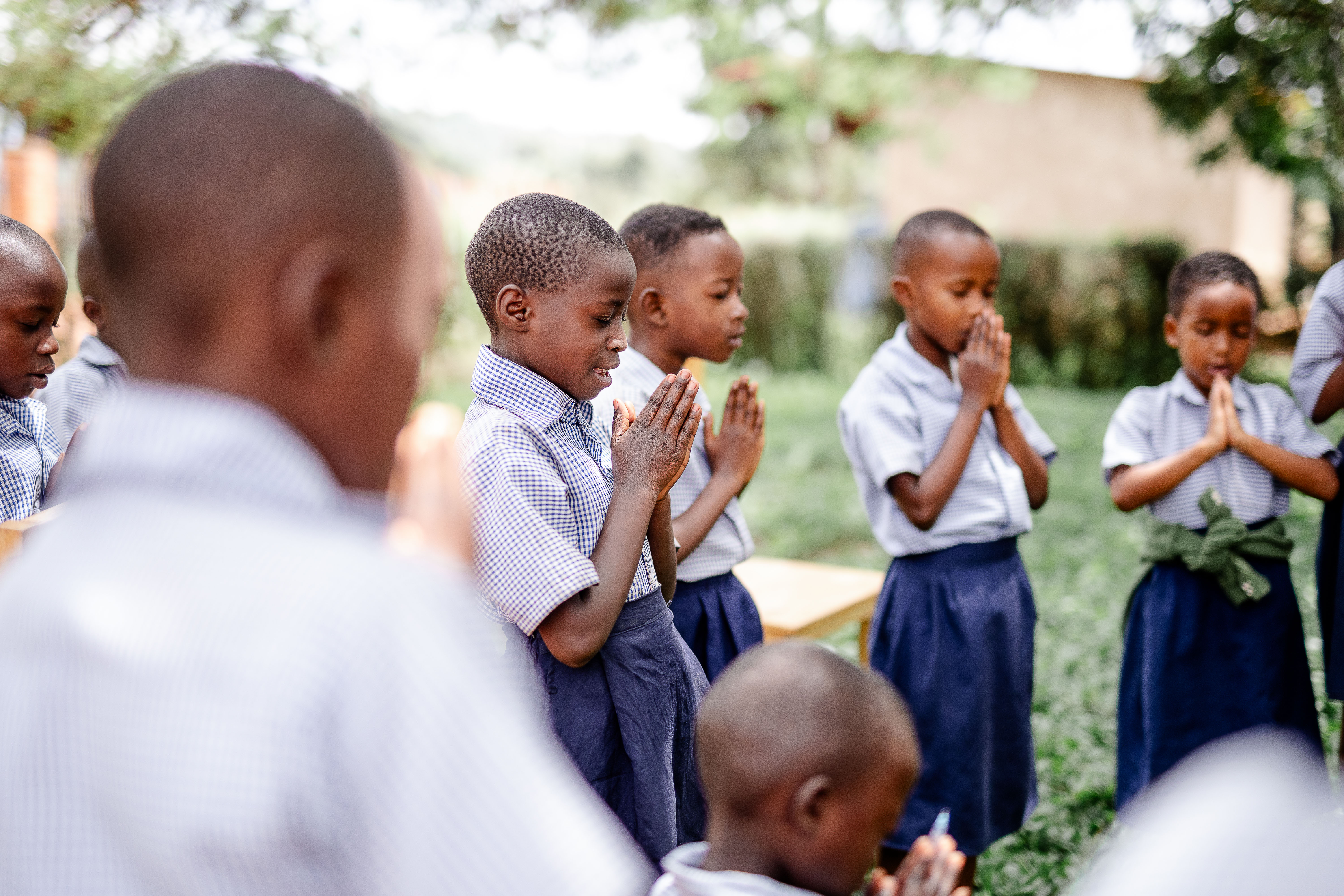  A group of children wearing school uniforms stand in a half circle and pray with their eyes closed and hands together in front of their chests.