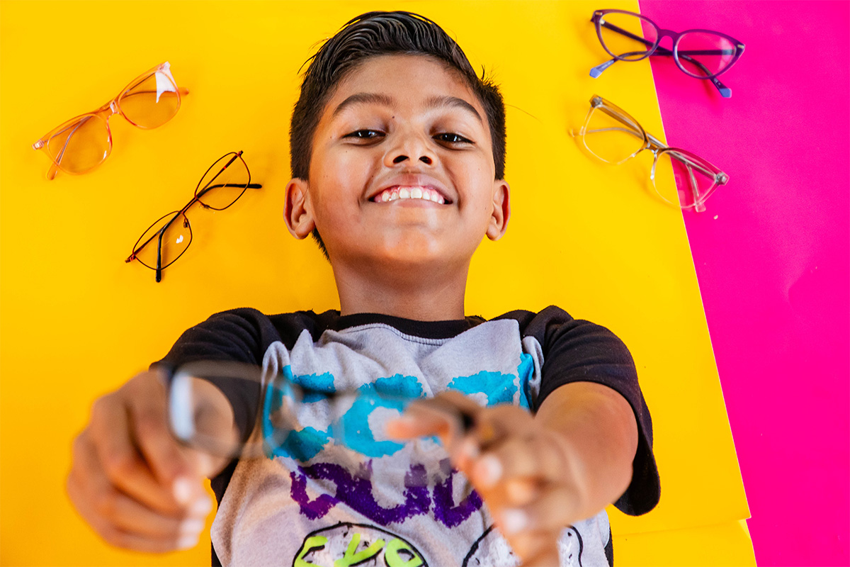 Young boy lies on a yellow and pink background and holds up glasses to the camera. He smiles brightly.