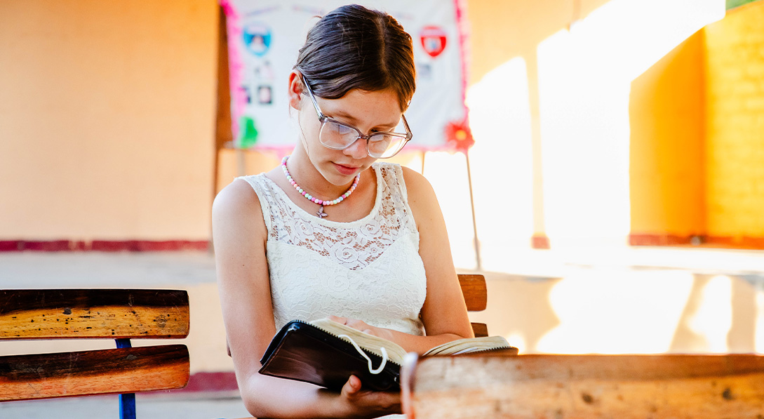 Young girl wearing glasses sits at a desk reading a Bible.