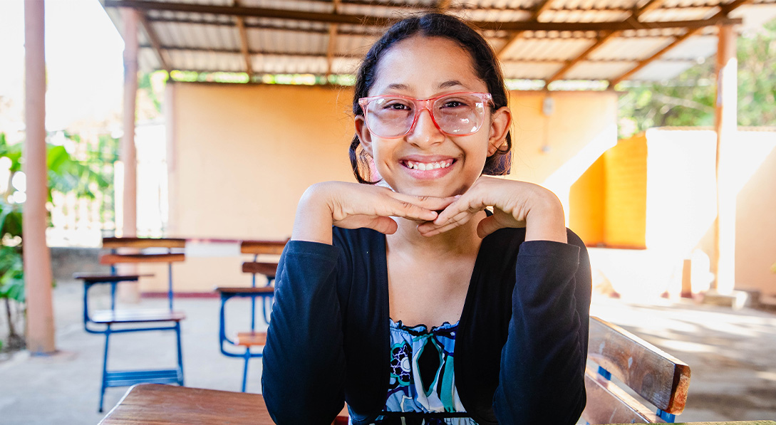 Young girl wearing pink glasses sits at a green table with her hands under her chin and smiles for the camera.