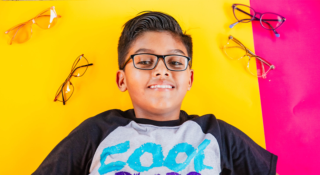 Young boy with glasses lies on a yellow and pink background. He smiles brightly.