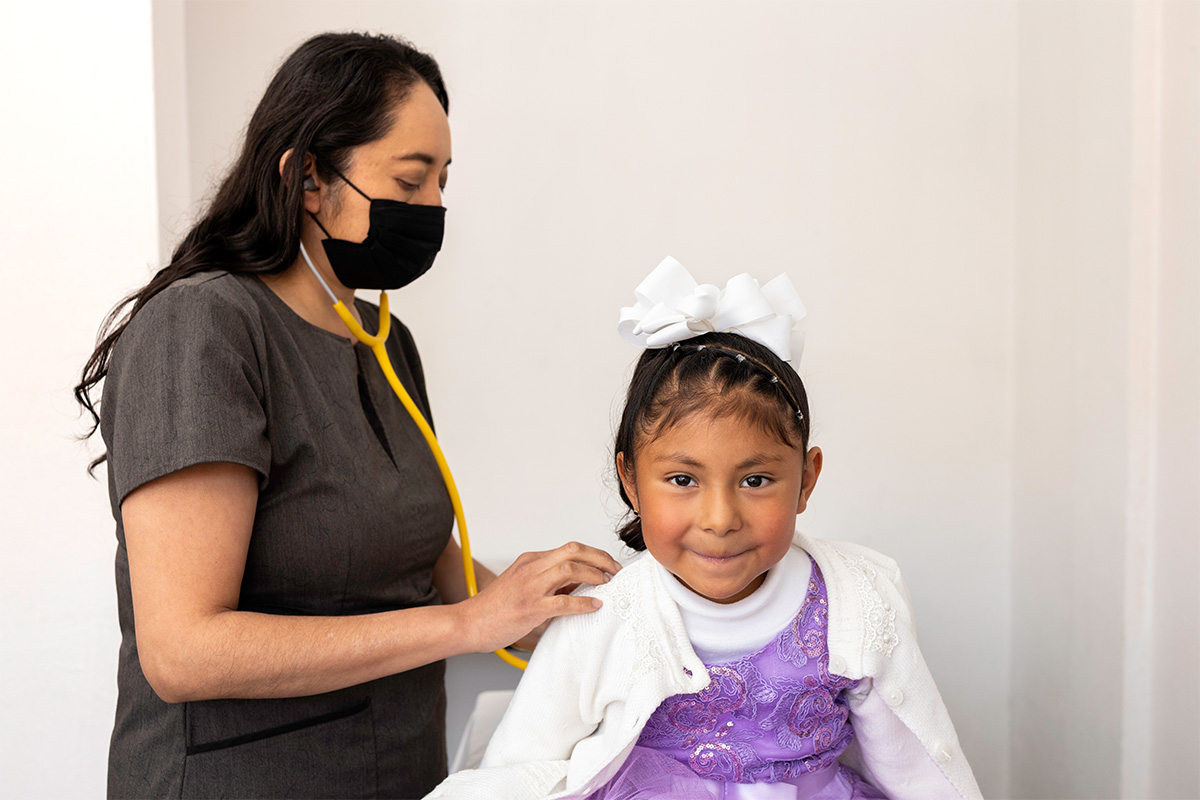 A young girl wearing a white bow smiles for the camera as a female doctor uses a stethoscope to listen to her breathing.