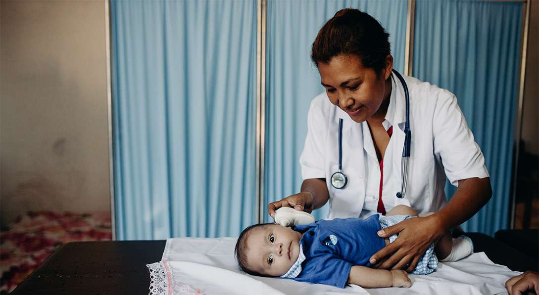 A baby boy wearing a blue shirt lies on a table as a female doctor take his temperature.