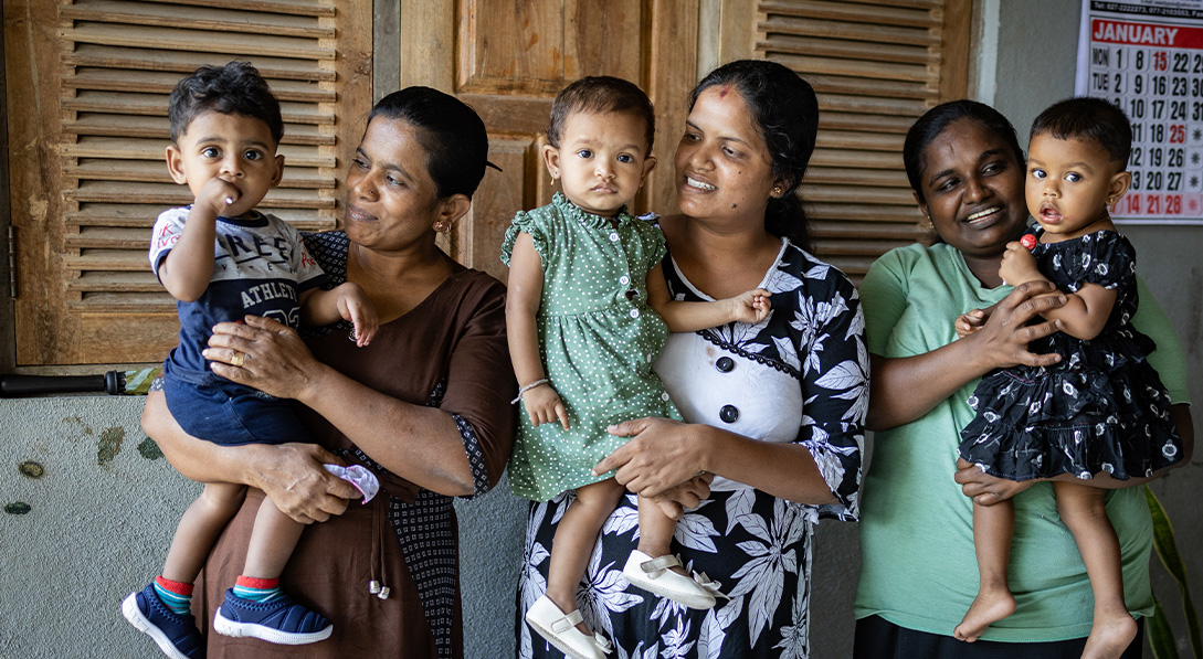 Three young children, one boy and two girls, are held by their mothers in front of wooden windows.