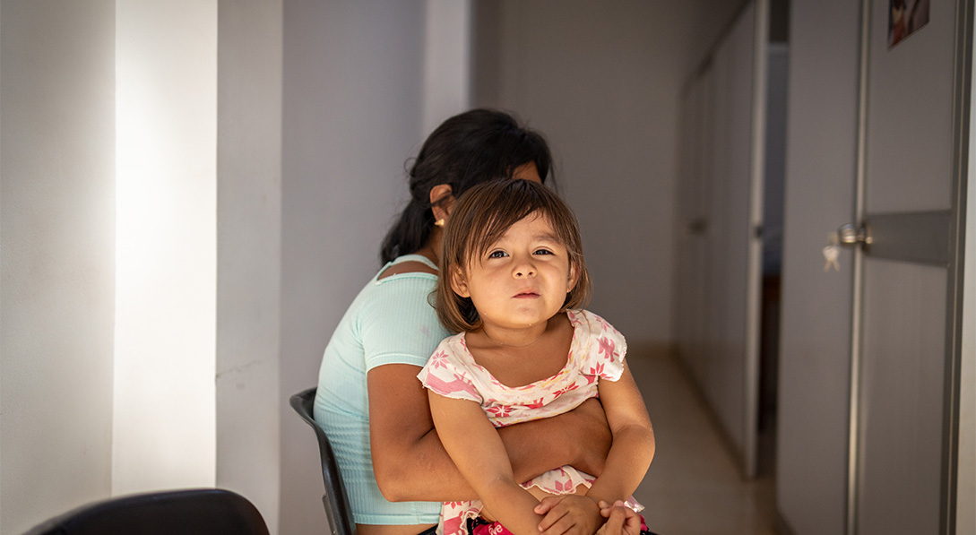 A young girl is held by her sitting mother while she smirks for the camera.