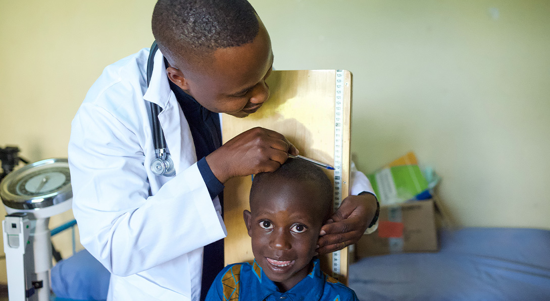 A young boy smiles for the camera as his male doctor measures his height.