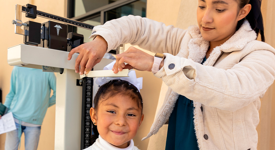 A young girl wearing a bright purple skirt smiles for the camera while a female nurse measures her height.