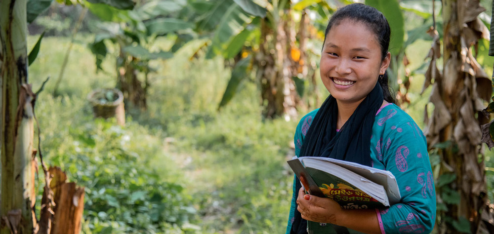 A smiling Shanti stands holding books and papers in one arm