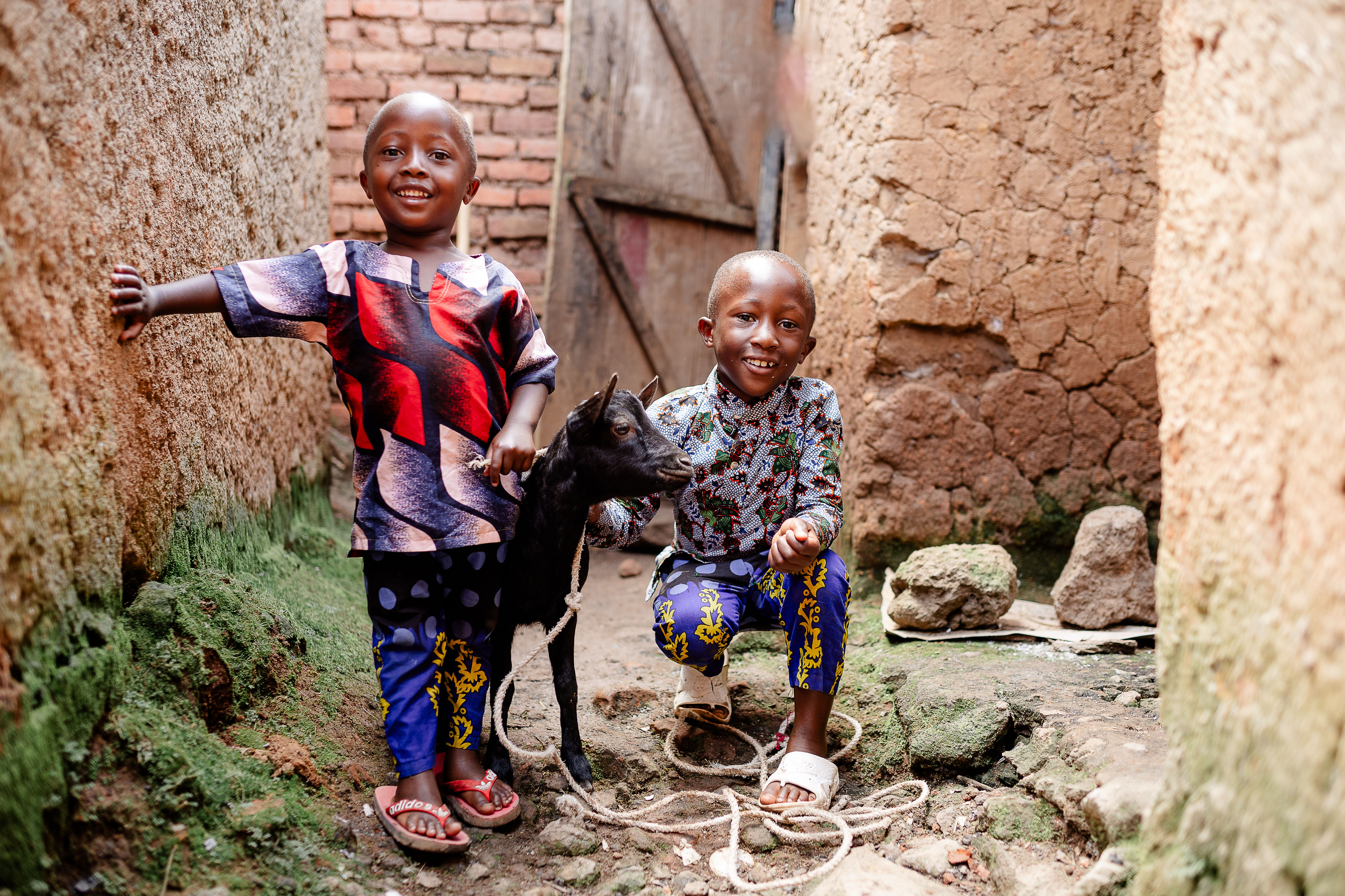  Two young boys stand in an alley smiling for the camera while petting a small black goat.