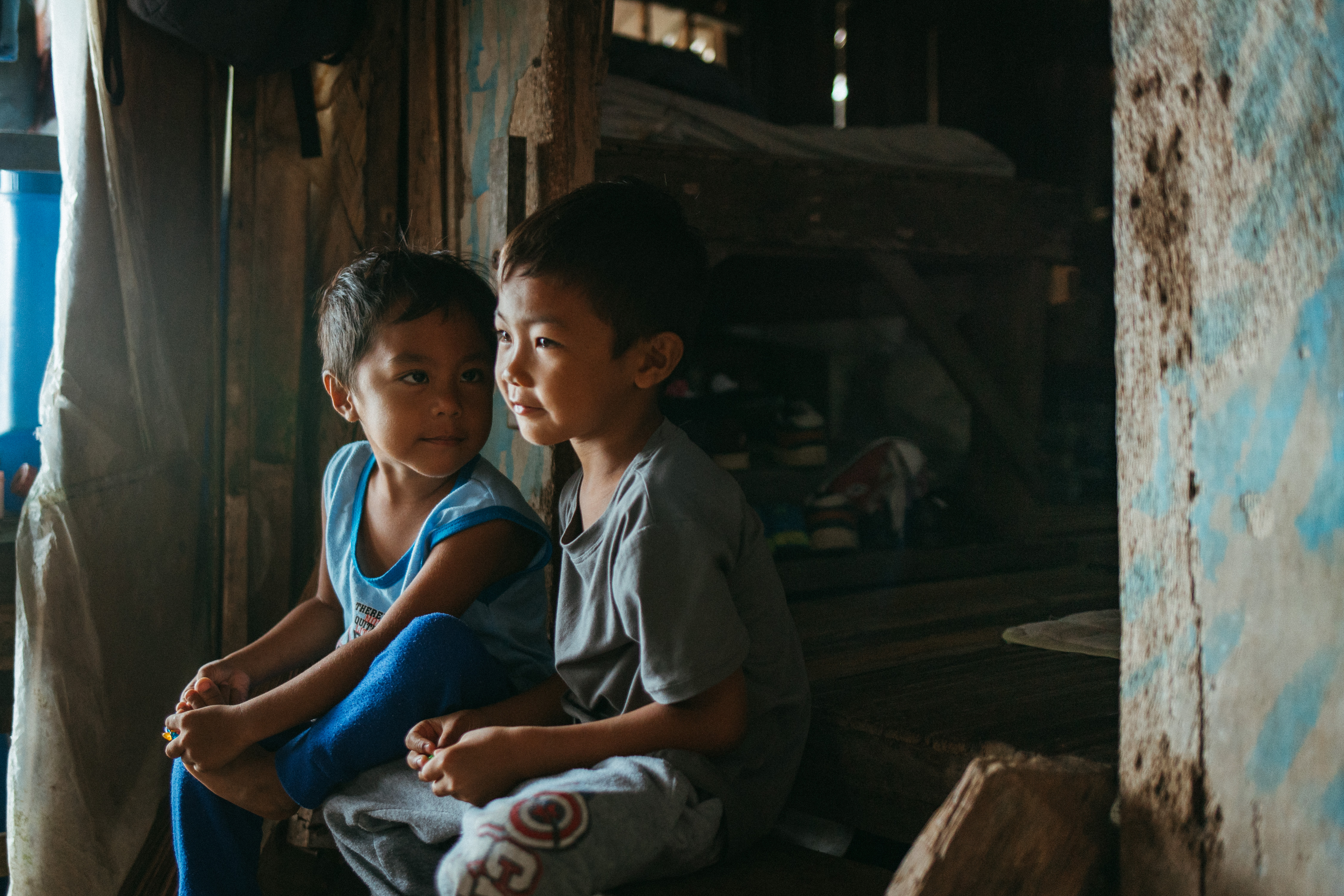 Two young boys sit next to each other inside their home.