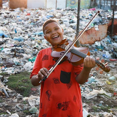 A boy wearing a red shirt stands in front of a trash-filled outdoor area. He is holding a violin and smiling.