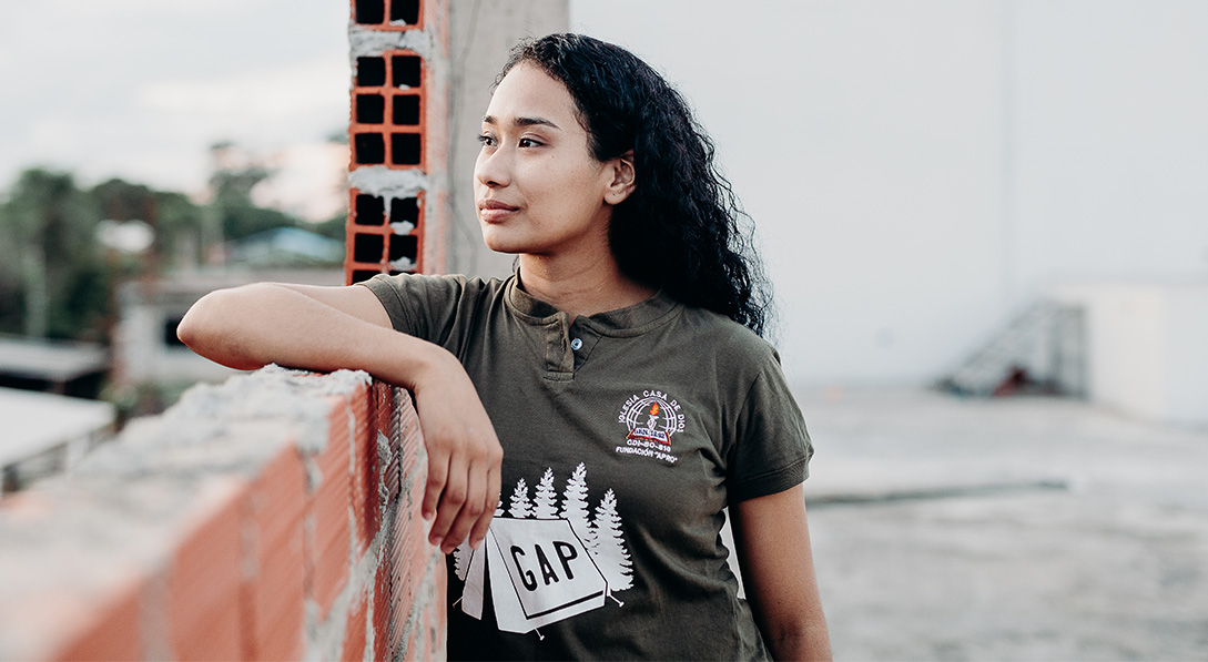 A young woman wearing a dark green shirt leans against an unfinished brick wall. She gazes into the distance thoughtfully.