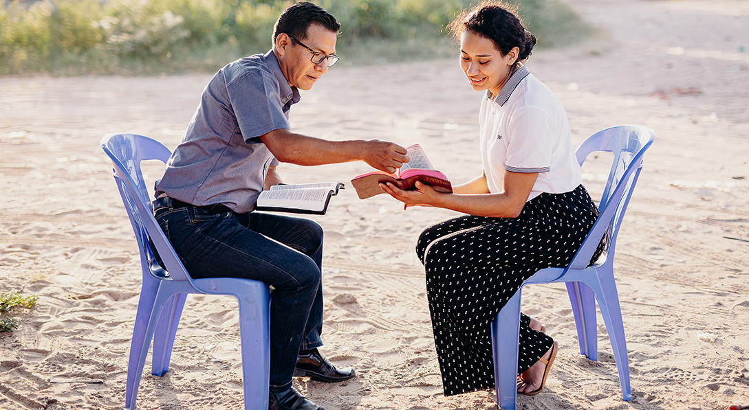 A man and a young woman sit across from each other on purple chairs outside. They both hold Bibles open, and the man points to a page in the woman’s Bible.