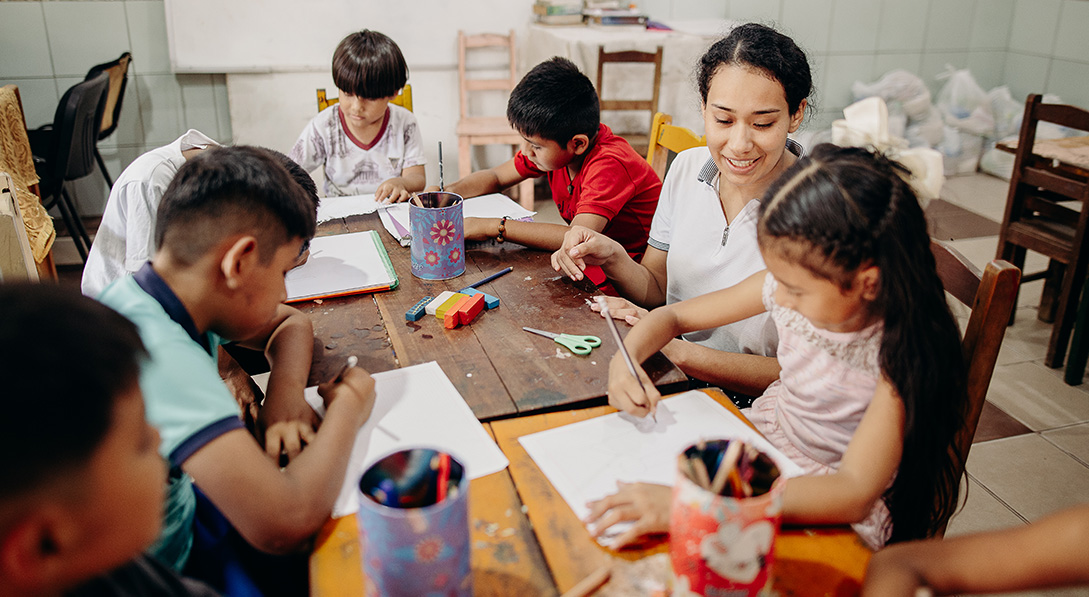 A young woman sits at a table with seven children as they practice handwriting on sheets of white paper. She is smiling at a little girl next to her.