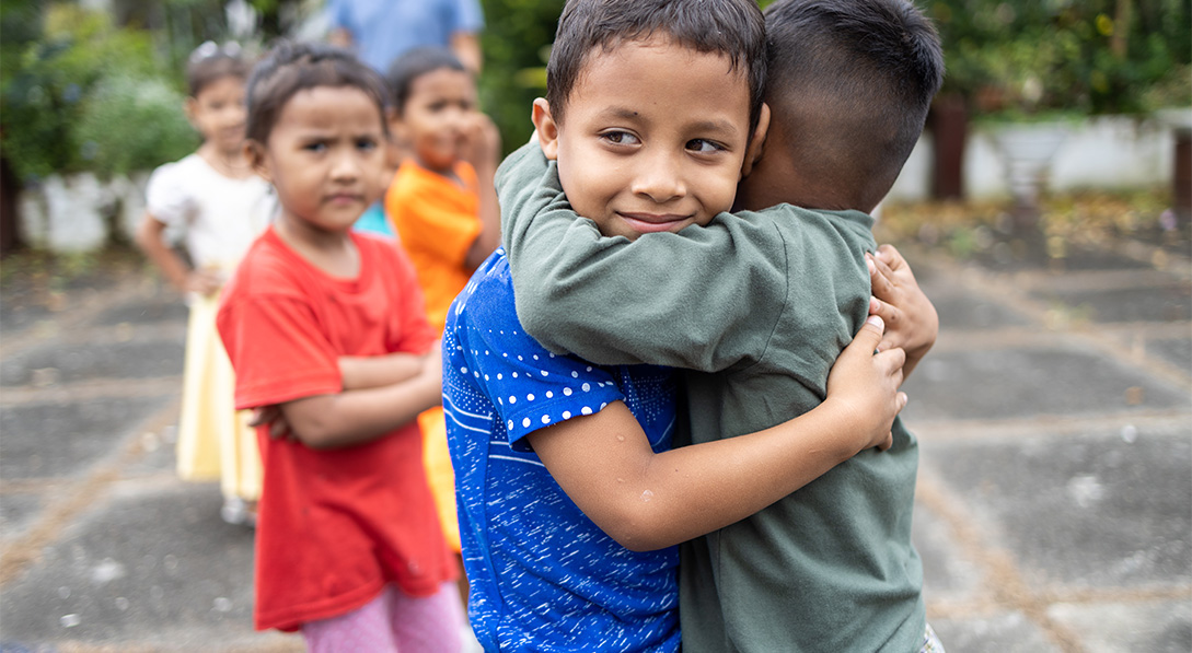 Two boys, one in a blue shirt and one in a green shirt hug each other on the playground.