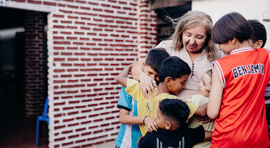 Sister Graciela gives a group hug to six young children outside of a brick building. She smiles.