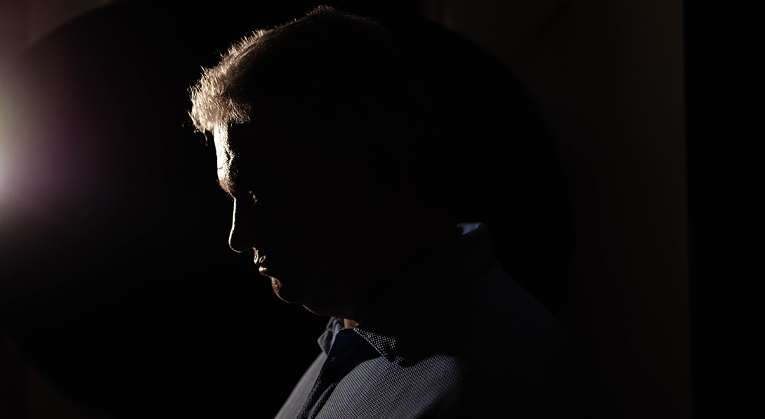 A silhouette of a man against a black background with light shining from the left just touching his face.