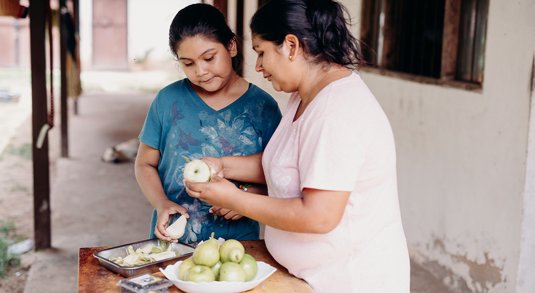 A girl and her mother stand at an outdoor table and prepare fruit for eating.