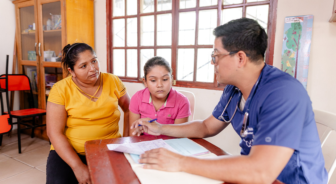 A young girl sits next to her mother at a desk and listens to a doctor.