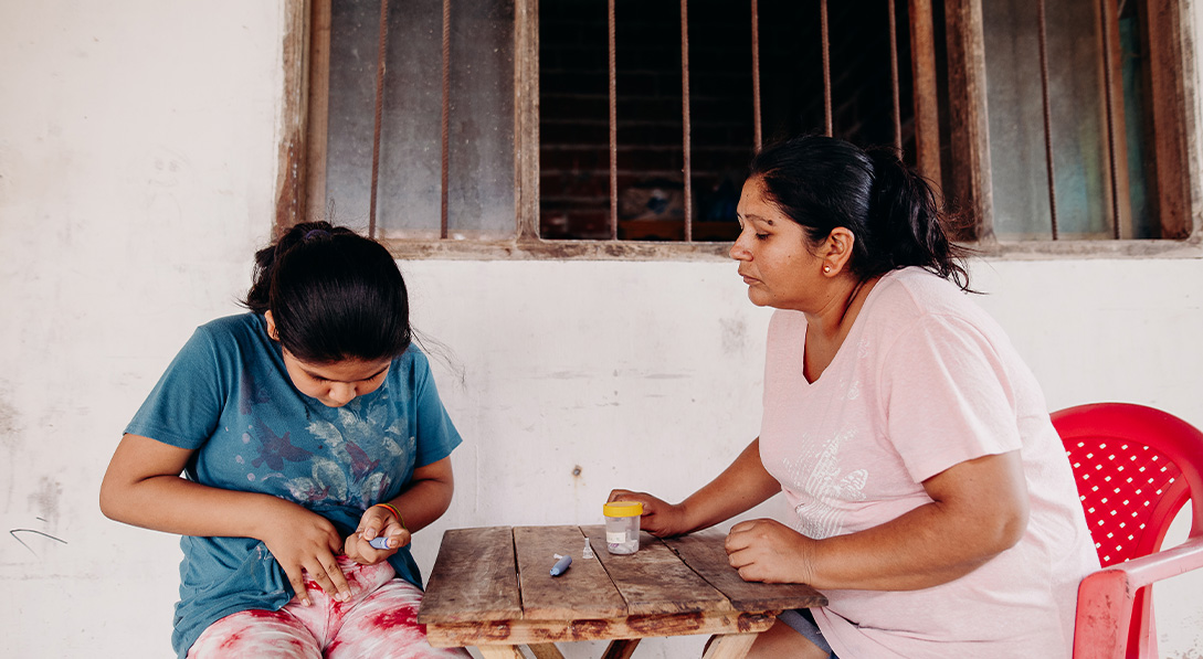 A mother watches from across a small table as her daughter gives herself an insulin shot in the stomach.