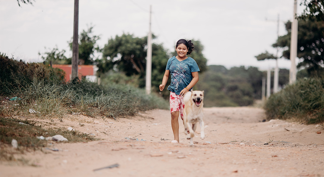 A girl smiles and skips down a dirt road while her dog runs in front of her.