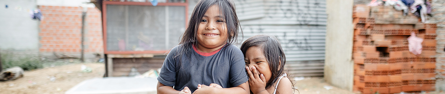 Two young girls stand in front of their home, smiling and giggling for the camera.