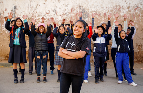 a girl with her arms crossed stands in front of a group of children with their hands in the air