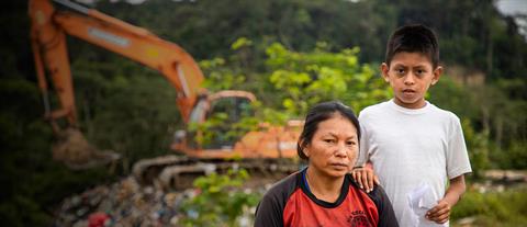 A young boy stands with his mother near a dilapidated home and looks at the camera.