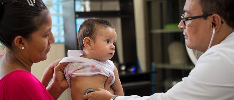 A doctor places his stethoscope on the belly of baby being held by its mother