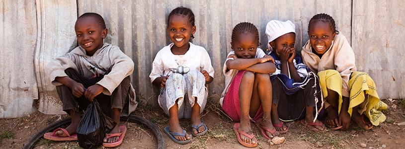 A group of children sitting on the ground with their backs to a corrugated metal well