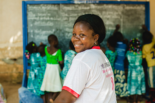 A teacher looks over her shoulder as students write on a chalkboard in the background