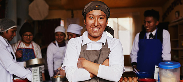 A young woman in an apron stands with her arms crossed and her hands tucked into her armpits